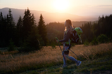 Woman hiker hiking outdoors at sunset. Sporty, positive woman traveling in mountains. Young, sporty female with backpack using trekking sticks, walking. Concept of harmony with nature.