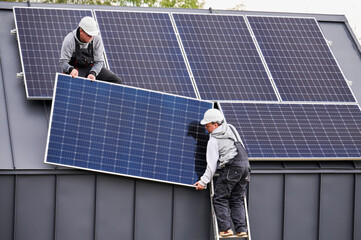 Men electricians installing solar panel system on roof of house. Workers in helmets lifting up...