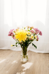 A bouquet of yellow and pink asters and gerbers in a glass vase against the backdrop of a window with rays of sun. Flowers in a vase in the interior