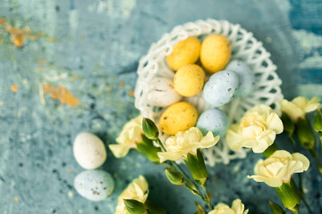 Fototapeta na wymiar Yellow beautiful flowers and blue, yellow colored easter eggs in wicker basket on a blue picturesque oil painting background close up. spring Mockups. Layout. Easter pastel contemporary background