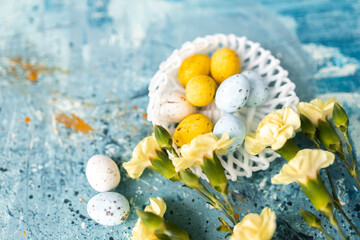 Obraz na płótnie Canvas Yellow beautiful flowers and blue, yellow colored easter eggs in wicker basket on a blue picturesque oil painting background close up. spring Mockups. Layout. Easter pastel contemporary background