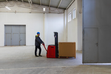 Young man in helmet, worker in warehouse transporting first palette through door to empty hall. Stock of Parcels with Products, transportation concept