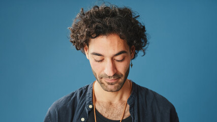 Close-up, man with curly hair, dressed in blue shirt, standing with his head down, isolated on blue...