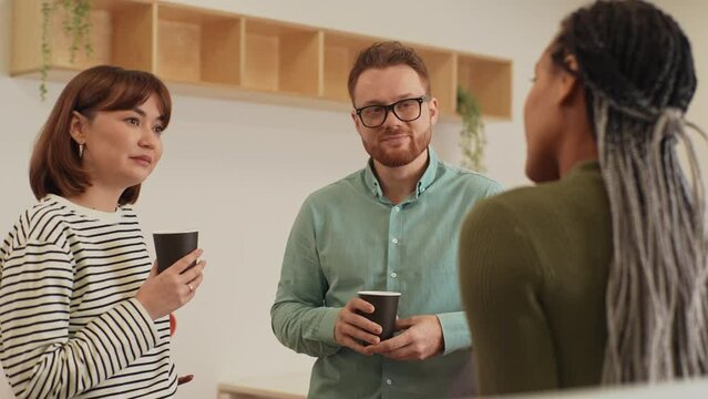 Three friendly multiethnic coworkers standing in office, drinking coffee and talking cheerfully in workspace during break. Male and female colleagues having conversation at modern open space.