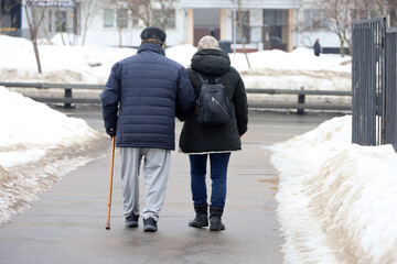 Elderly man with cane and woman walking on city street, rear view. Old couple in warm clothes during snow weather
