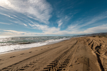 Fototapeta na wymiar Expansive Sandy Shoreline with Tire Tracks Stretching Towards the Horizon Under Blue Skies