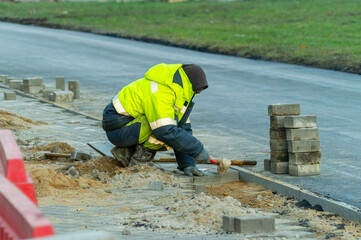 A man in a yellow jacket is laying bricks on the street. The bricks are stacked in a row