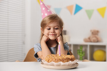 Cute girl in party hat with birthday cake at table indoors