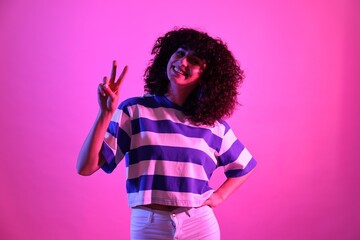 Beautiful young woman showing V-sign on color background in neon lights