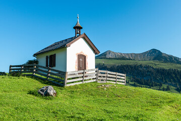 Kapelle mit weisser Fassade auf einer Alm in den Tiroler Bergen im Sonnenlicht vor blauem Himmel,...