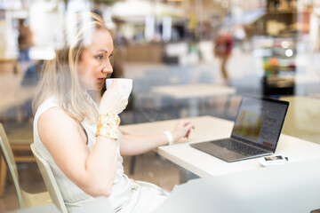 Business woman drinks coffee and works on laptop while sitting at table in cafe. View through window