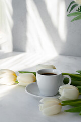 Fototapeta na wymiar Hot coffee drink cup and white tulip flowers bouquet on table with bright natural sunlight pattern on wall and table, good morning concept, aesthetic business branding template