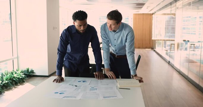 Two millennial multinational businessmen discuss financial report in charts, diagrams, graphs, make sales statistics analysis, review deal benefits at meeting in modern board room. Teamwork. Finances