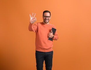 Portrait of smiling young businessman with digital tablet showing OK sign over orange background