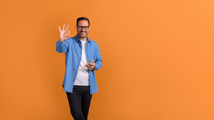 Portrait of smiling male professional using mobile phone and showing OK sign over orange background