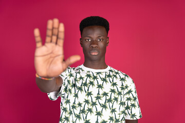 Young African man making the stop gesture with his hand wearing a summer t-shirt with drawings of palm trees on a crimson red background with copy space. Stop racism concept.