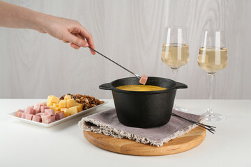 Dipping piece of ham into fondue pot with tasty melted cheese at white table, closeup