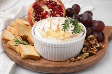 Board with tasty baked camembert, croutons, grapes, walnuts and pomegranate on white tiled table, closeup
