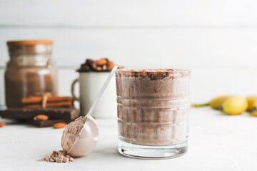 Glass with chocolate protein drink, milkshake smoothie on white table with bananas, protein powder in measuring spoon, protein bar, almond nuts and cinnamon sticks
