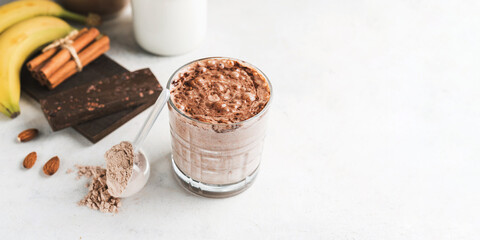 Glass with chocolate protein drink, milkshake smoothie on white table with bananas, protein powder in measuring spoon, protein bar, almond nuts and cinnamon sticks