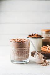 Glass with chocolate protein drink, milkshake smoothie on white table with protein powder in measuring spoon, protein bar, almond nuts and cinnamon sticks