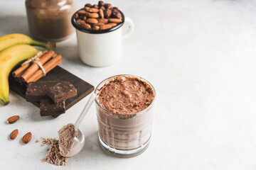Glass with chocolate protein drink, milkshake smoothie on white table with bananas, protein powder in measuring spoon, protein bar, almond nuts and cinnamon sticks