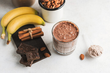 Glass with chocolate protein drink, milkshake smoothie on white table with bananas, protein powder...