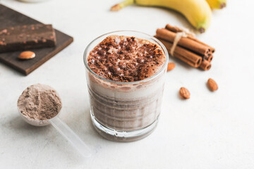 Glass with chocolate protein drink, milkshake smoothie on white table with bananas, protein powder in measuring spoon, protein bar, almond nuts and cinnamon sticks