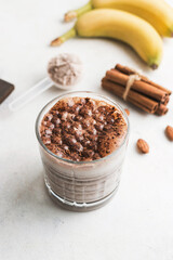 Glass with chocolate protein drink, milkshake smoothie on white table with bananas, protein powder in measuring spoon, protein bar, almond nuts and cinnamon sticks