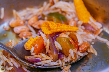 A spoon filled with lots of different foods, rice, pieces of cooked onions and roasted peppers, peanuts and a corn cone chip. Selective focus on foreground with a full metal plate in  blur background 