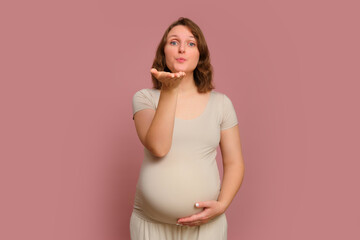 A pregnant woman blows a kiss on a studio pink background. Pregnancy in a woman with a belly, copy space