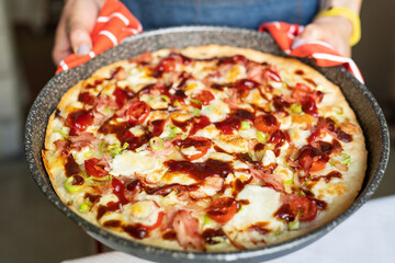 The girl is holding a freshly baked pizza with melted cheese, meat, leeks and toppings and wearing oven mitts.
