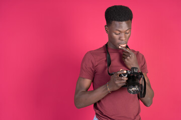 Young black man in red t-shirt with a mirrorless camera in his hands looking at the screen and checking the photo with a thoughtful face with a carmine red background and copy space.