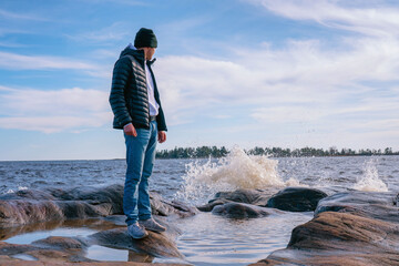 Handsome young men in jacket, jeans and sport shoes stands on wet stone coastline to calm sea and...