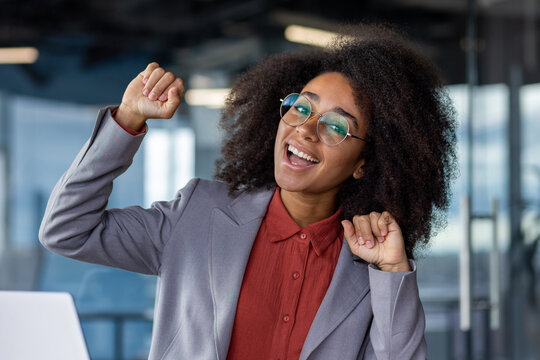 Close-up photo of a young African-American woman in the office, sitting at the workplace and rejoicing in success, raising her hands up and looking at the camera.