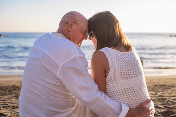 Rear view of bonding senior couple hugging while sitting on the beach face the sunset enjoying vacation and retirement, two elderly people expressing love and tenderness