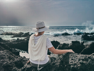 Back view of senior woman sitting on rocky beach watching ocean waves crashing with white foam....