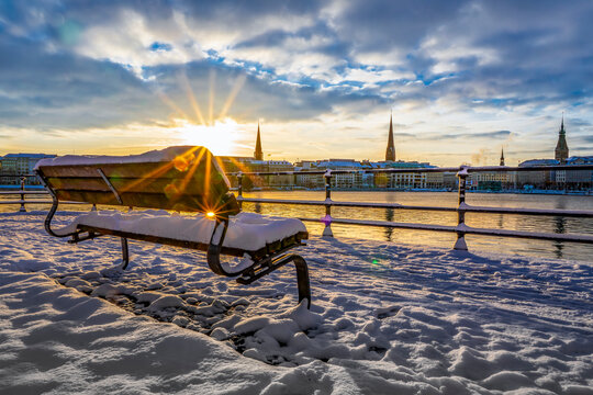 Verschneite Parkbank am Ballindamm in Hamburg morgens im Winter bei Sonnenaufgang, im Hintergrund unscharf die Geb&auml;ude an der Alster zu sehen, horizontal