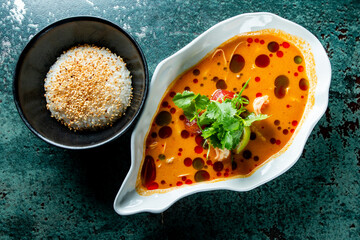 Top view of a gourmet spicy shrimp soup garnished with herbs, served with a side of sesame sprinkled white rice on a textured surface