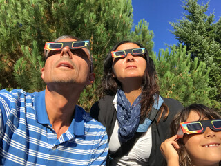 Father, mother and daughter, family viewing solar eclipse with special glasses in a park - obrazy, fototapety, plakaty