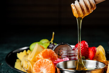 Vibrant fresh fruits and cheese on a plate, with golden honey drizzling from a wooden dipper, against a dark background