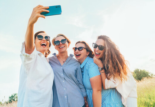 Portrait of four cheerful smiling women in sunglasses embracing together and making selfie photo using modern smartphone during outdoor walk. Woman's friendship, relations, and happiness concept image