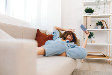 Happy woman sitting on a cozy sofa, holding a mobile phone and smiling while enjoying online games, reading, and taking a selfie at home