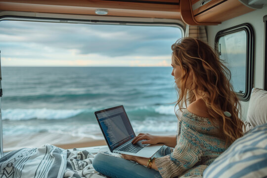 A woman sitting in an RV working on laptop. The background is the ocean