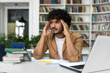 An anxious graduate student surrounded by books at a library desk, studying hard and preparing for exams on campus, symbolic of academic pressure and university admissions preparation.
