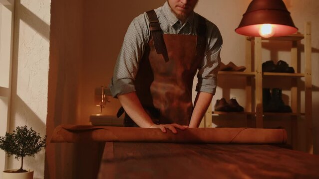 Shoemaker rolls out piece of leather on table in workshop. Artisan works with leather to make handmade shoes.