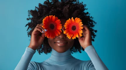 Fototapeten A person holds bright orange gerbera flowers over their eyes like glasses, smiling broadly against a blue background, creating a playful and joyful portrait. © MP Studio