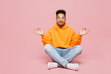 Full body spiritual young man of African American ethnicity wear yellow hoody casual clothes sit hold spread hand in yoga om aum gesture relax meditate try calm down isolated on plain pink background - obrazy, fototapety, plakaty