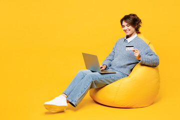Full body young IT woman in grey sweater casual clothes sit in bag chair using laptop pc computer...