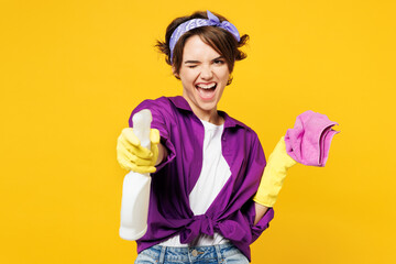 Young woman wearing purple shirt rubber gloves do housework tidy up hold in hand rag spray bottle look camera wink blink eye isolated on plain yellow background studio portrait. Housekeeping concept.
